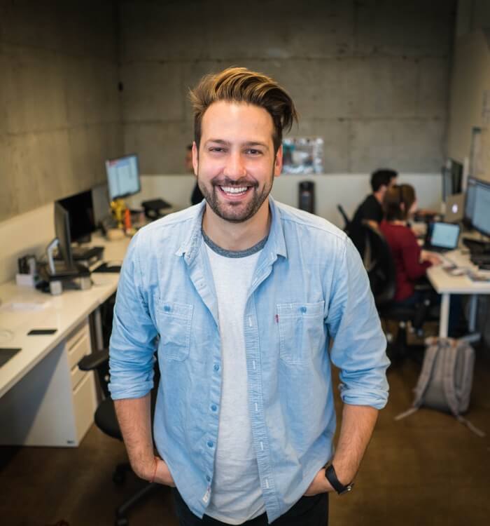 Team1 Photographic image depicting a young man wearing business casual attire. The man smiles. Behind him are several people working at computer stations.