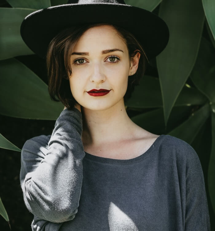 Team2 Photographic image of a young woman standing among several large tropical leaves. The woman wears an eccentric hat, and a loose gray shirt.