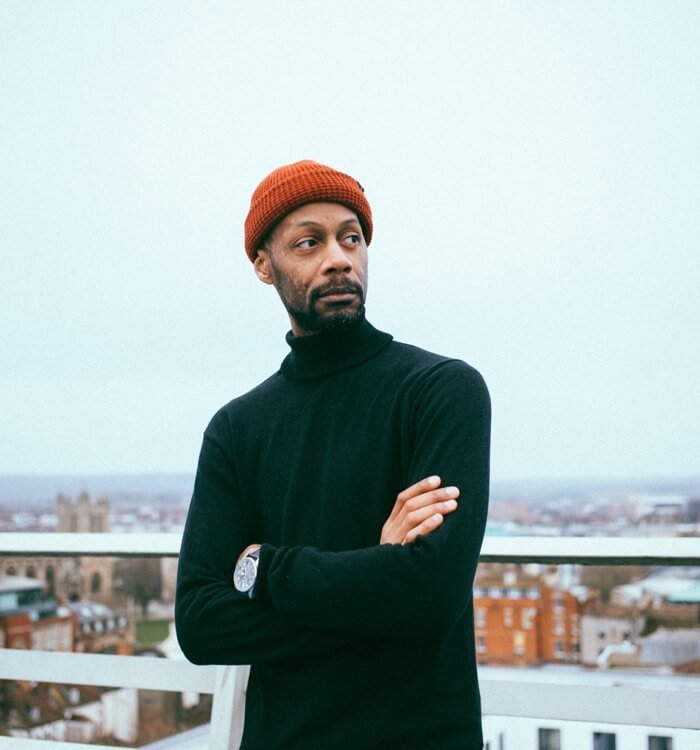 Team3 Photographic image depicting a lean man wearing a brick-colored wool cap. The man leans against a white metal railing, with a blurred township visible in the background.