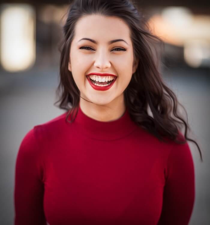 Team6 Photographic image of a young woman with dark hair against a blurred background. The women wears a red sweater, and bright red lips stick.