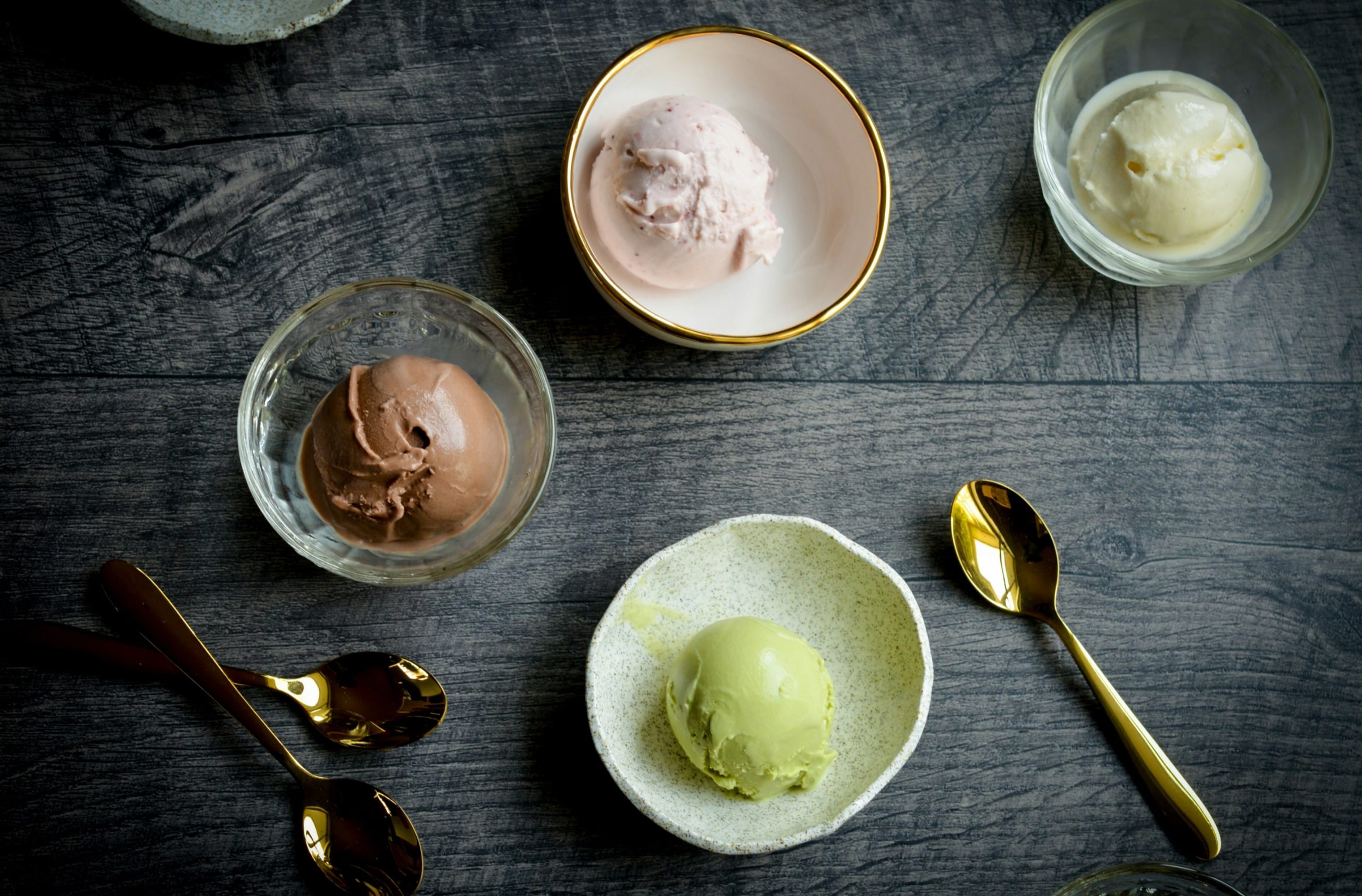 Photographic image depicting a wooden table, upon which sits four different bowls. Each bowl contains a scoop of different types of ice-cream - Vanilla, Strawberry, Chocolate, and Pistachio