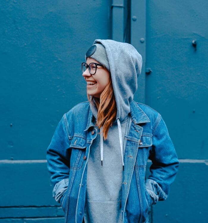 team4 Photographic image depicting a young woman laughing against a blue background. The woman wears a denim jacket and a gray hoodie.