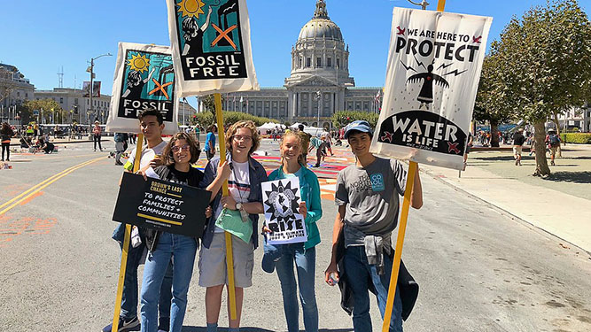 Five young individuals protesting outside their state capital, holding signs advocating for a fossil-free future.