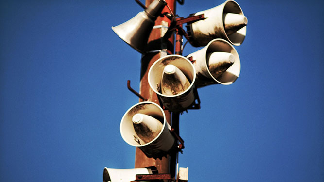 A wooden pole adorned with several white loudspeakers pointing in every direction, in front of a blue sky.