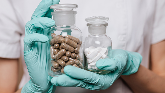 A person in a white uniform and green latex gloves holds two glass jars containing white pills and capsules filled with grey herbs.