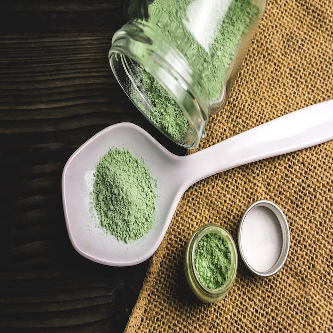 Photographic image depicting a woven placemat on a wooden table. On top of the mat, are two glass jars filled with green kratom powder, as well as a white ceramic spoon filled with a measure of kratom powder.