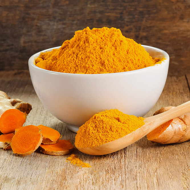 Orange Turmeric Powder in a white bowl with a wooden spoon and tumeric roots