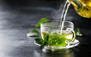 Image of a Glass Tea Cup containing several bright green tea leaves, being filled with boiling water.