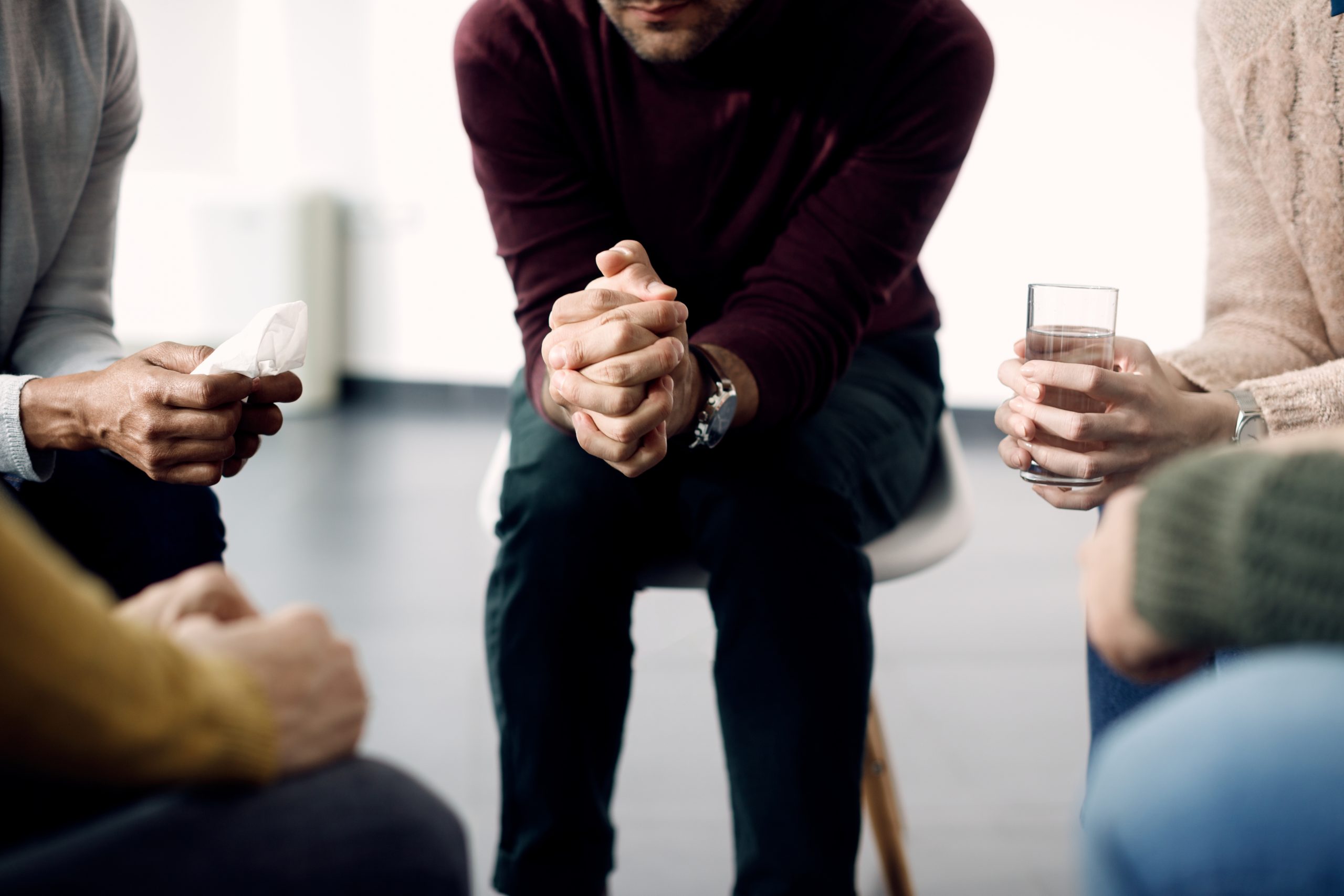 Photographic image depicting a close up of a bunch of people sitting in a circle. The people's faces aren't visible, but the central figure is leaned forward clasping his hands as if he's about to engage in a serious discussion of Opioid Advocacy