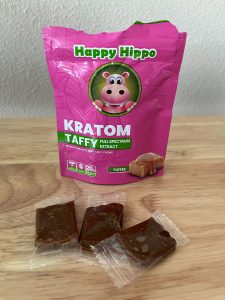 A packet of Happy Hippo brand, Kratom Extract (Toffee Flavored) Taffy Chews, with the top of the package ripped open and a few individually wrapped taffy chews lying before it.